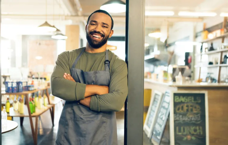 Homem sorridente de avental em frente ao seu café, representando empreendedorismo e atendimento acolhedor