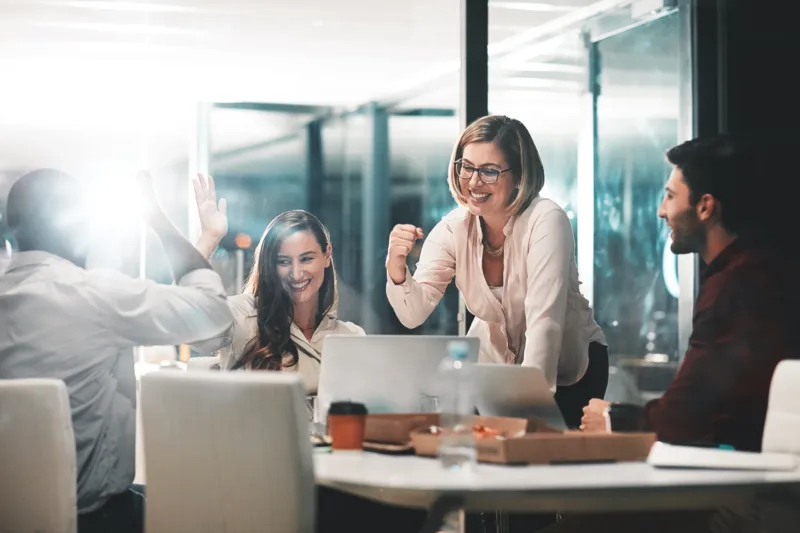Equipe de profissionais celebrando resultados em reunião noturna, com laptops e pizza sobre a mesa